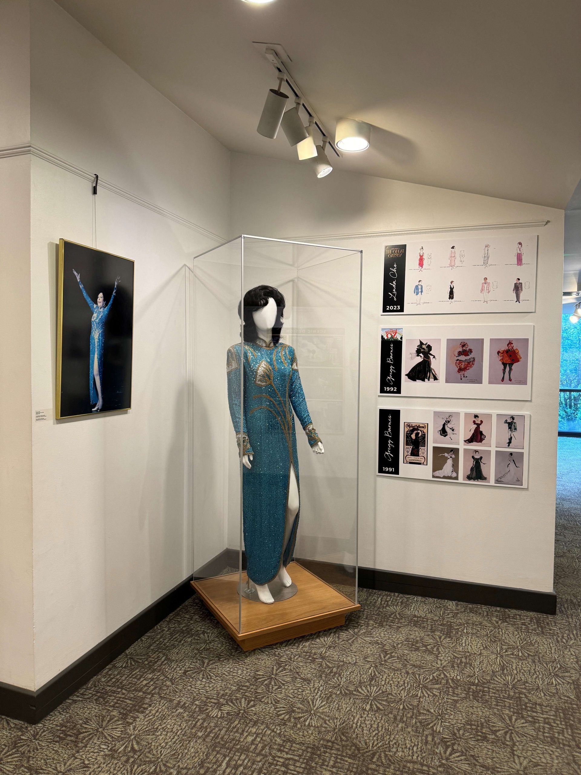 Mannequin wearing a blue and silver sparkle dress in a case. A picture on the left wall is of Anne Miller wearing the dress. Pictures on the right wall show artist costume sketches for several shows.