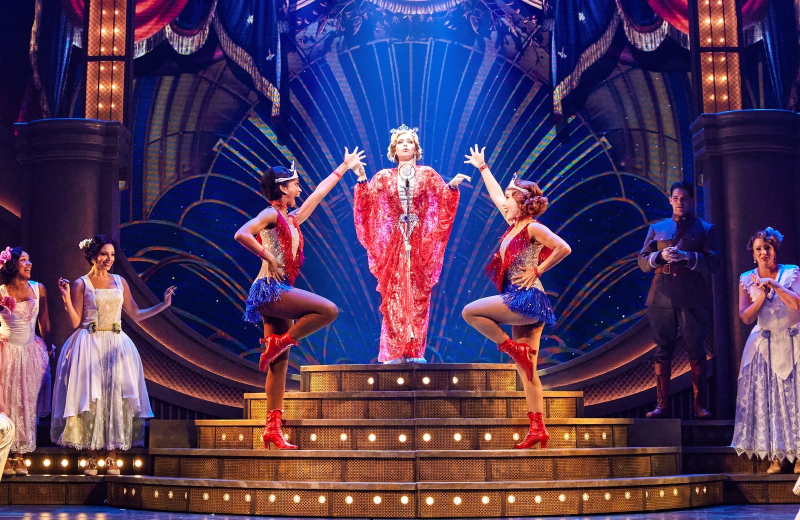 Woman in a red dress in front of a microphone on an elevated stage. Two women on either side with rasied hands. Other actors are on both sides of the stage. The background is blue with touches of gold.