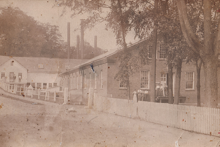 Diamond Paper Mill 1900 - white building in the background with four smoke stacks, large brick building the front all behind a fence with mill workers by the doors