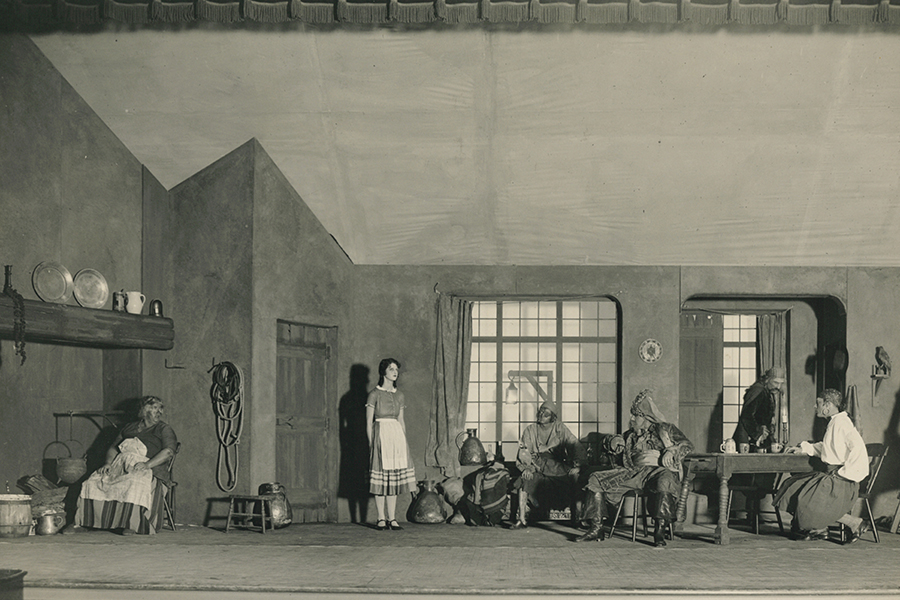 Newark Arts Theatre - left to right a woman seated by a fire, girl standing by a door, men seated at tables - all facing the girl