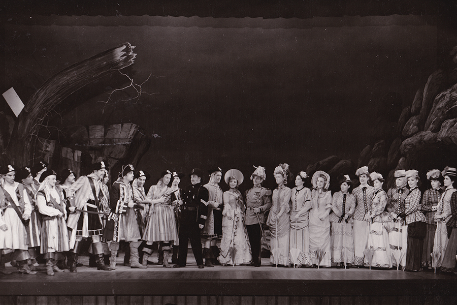 Pirates of Penzance - pirates on the left, a policeman in the middle and townspeople mostly women on the right against a background of rocks and trees