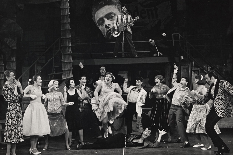 Grease 70s - a group of actors playing high school students and some teachers in 50's attire dancing in a semi-circle, one man is lying down while a woman jumps over him. In background is a man with a guitar standing in front of a large headshot of James Dean