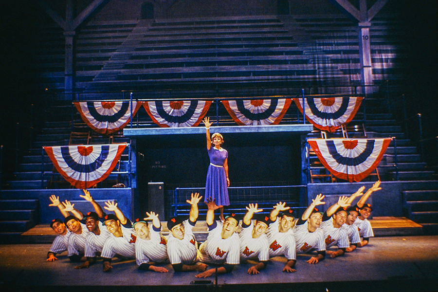 Damn Yankees 80s - Lola stands in the center with her right arm outstretched surrounded by baseball players in a semi-circle at her feet also with outstretched arms