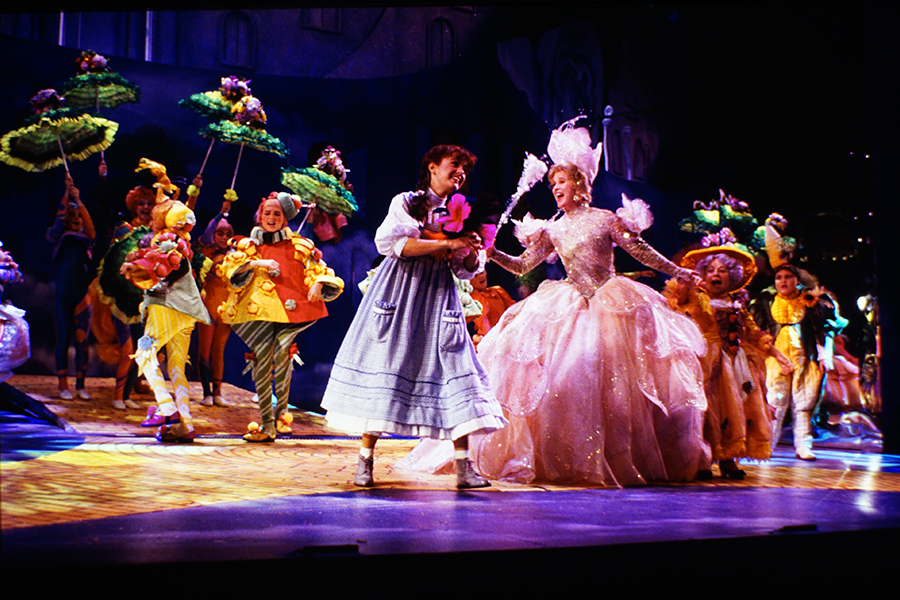 Wizard of Oz 90s - munchkins dancing behind Dorothy and Glinda in front on the yellow brick road